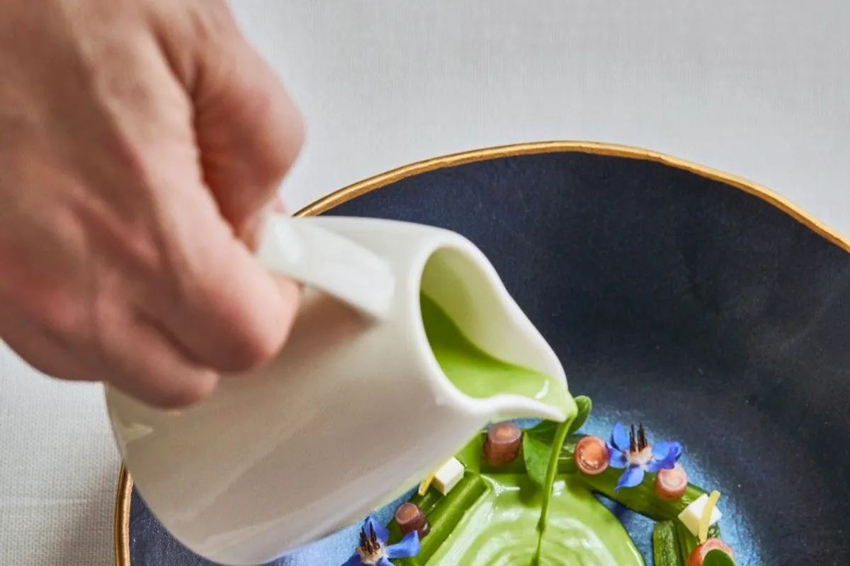 Plato con crema verde, verduras y flores comestibles mientras se vierte una salsa